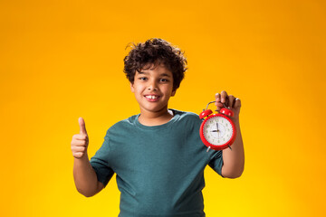 Child boy holding alarm clock and showing thumb up gesture  over yellow background. Time management...