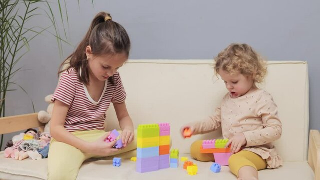 Playful girl with building toys. Colorful blocks for creative play. Adorable little girls playing with colorful toy blocks together while sitting on couch at home in living room interior