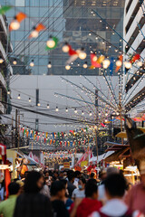 A vibrant market street in Bangkok under a canopy of colorful lights and flags, bustling with...