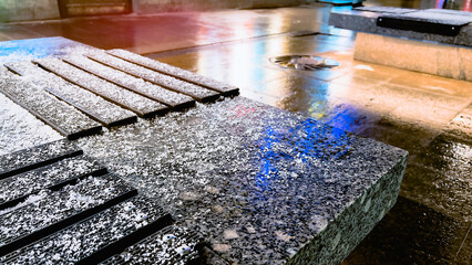 Stone bench against wet asphalt road. Glowing light reflections on tiled pavement at night. City...