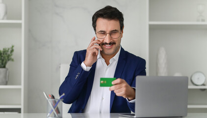 Smiling businessman in a blue suit talking on the phone and holding a green credit card
