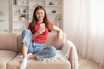 Relaxed young asian woman sitting on couch and looking at her smartphone