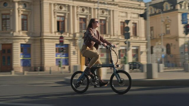 Stylish young woman riding an electric bicycle in city centre in modern street during fall season. Concept of urban cycling and eco transportation by bike.
4k slow motion with copy space