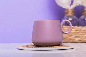 A purple mug on a fabric coaster, set on a white table with white flowers and a purple background in the background. Ideal for cozy home decor or coffee shop ambiance.
