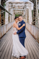 Valmiera, Latvia - July 7, 2023 - A bride and groom embrace on a rustic bridge, smiling and looking...