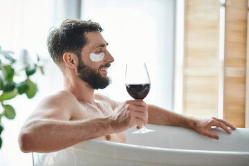 cheerful good looking man with beard and eye patches relaxing in bathtub with glass of red wine