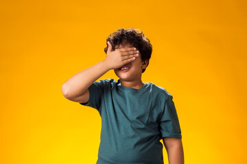 Upset kid boy coverihg eyes with hand over yellow background. Bad emotions and bulling concept