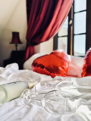 Two glasses and a bottle of champagne and heart-shaped balloons in blurry focus are on a white bed in a retro bedroom. Wedding day or Valentine’s Day.
