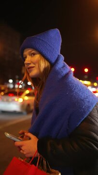 Smiling woman in blue hat with shopping bags uses her smartphone to call taxi on lively city street at night, surrounded by glow of urban lights. City shopping, evening leisure and transportation