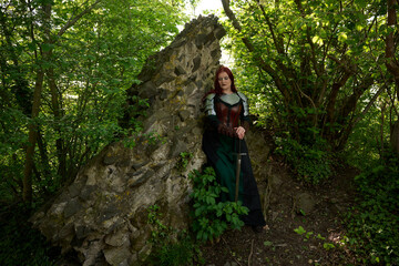 Mittelalterliche Frau mit Rüstung und roten Haaren als Kriegerin mit Kriegsbemalung mit Schwert im Sommer vor einer alten Burgruine, mit Korsett aus Leder und Umhang