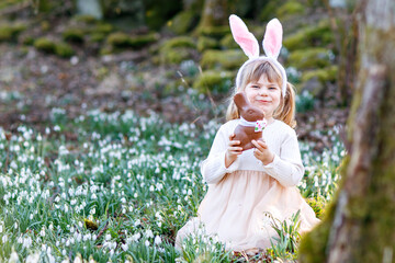 Happy little girl with Easter bunny ears eating chocolate figure in spring forest on sunny day,...