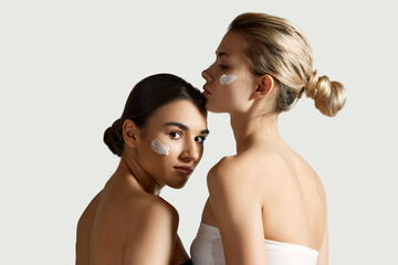 SPF face care. Brunette and blonde young girls with cream on cheeks against white studio...