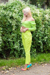 Full-length portrait of a beautiful young blonde with long hair in a yellow-green pantsuit and bright high-heeled pumps against a background of green bushes with flowers