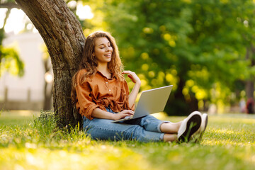 Portrait of amazing beautiful woman sitting on grass outdoors using laptop computer. Concept for...