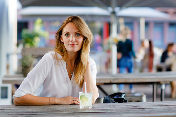 Young woman drinking mojito cocktail at cafe terrace at hot summer day. Beautiful businesswoman enjoying warm evening at afterwork restaurant. Happy smiling alone lady.
