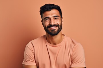 Portrait of handsome young bearded Indian man looking at camera and smiling, standing against brown background