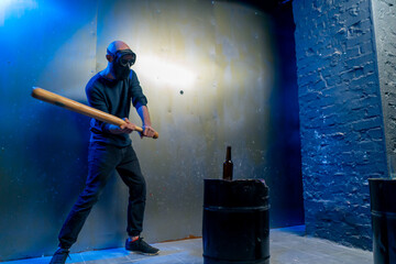 A man in a protective mask smashes a glass bottle with a bat in a crash room for stress relief