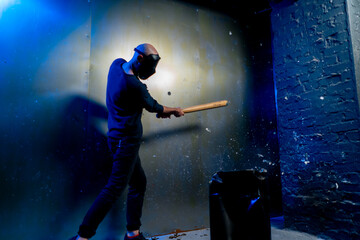 A man in a protective mask smashes a glass bottle with a bat in a crash room for stress relief