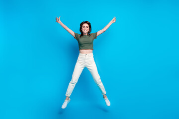 Fototapeta na wymiar Full size photo of good mood lovely woman wear green t-shirt white trousers jumping raising hands up isolated on blue color background