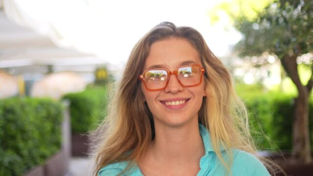 Smiling woman in trendy glasses walking on cafe terrace. Beautiful blond female in blue shirt looking happily into camera. Young lady on greenery and street garlands background.