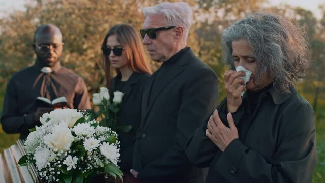 Side medium footage of Caucasian senior lady praying and crying standing with family by wooden casket with white flowers during memorial ceremony held by Black priest at cemetery