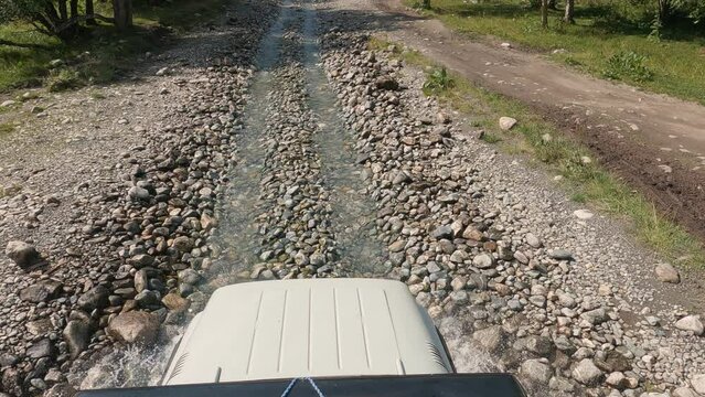 An SUV is driving along the bed of a shallow river in the mountains. First person view of the bonnet and the road. Off-road driving on a mountain road. Slow motion