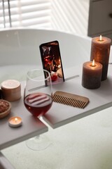 White wooden tray with smartphone, glass of wine and burning candles on bathtub in bathroom