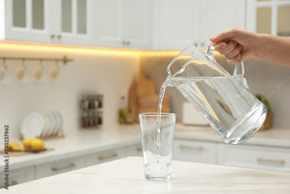 Poster Woman pouring water from jug into glass at white table in kitchen, closeup. Space for text