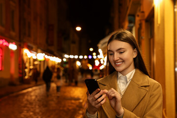 Smiling woman using smartphone on night city street. Space for text
