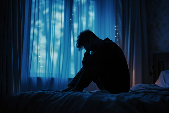 Silhouette of a guy sitting on a bed in front of a window he's upset sad, didn't get enough sleep