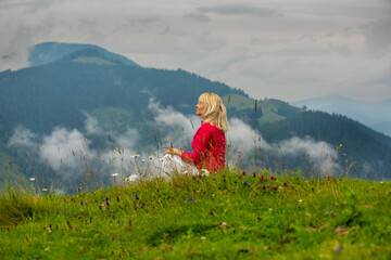 A middle-aged blonde woman sits in a clearing high in the mountains among wild flowers and clouds. A moment of pleasure.