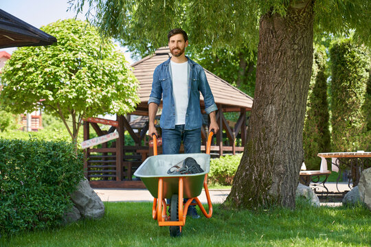 Caucasian bearded man pushing wheelbarrow, while working in garden in sunny day. Front view of smiling guy in denim shirt using cart for gardening, while looking at camera. Concept of seasonal work.