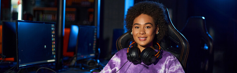 happy african american female gamer with headphones sitting on comfortable gaming chair, banner