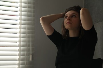 Sad young woman near closed blinds indoors, space for text