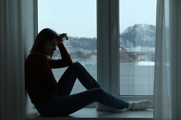 Sad young woman sitting on windowsill near window at home