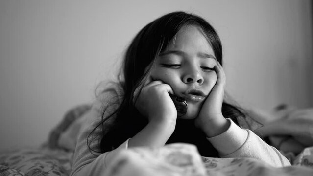 Sad depressed little girl laid in bed holding head with hands in chin, close-up face of melancholic child feeling down in black and white, monochrome