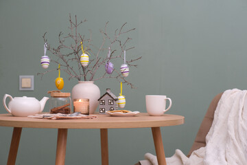 Beautiful Easter table setting with festive decor indoors. ceramic vase with tree branches and easter eggs, white teapot, chair with cover near the wooden table