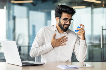 Overworked indian businessman feeling unwell at office desk with laptop and inhaler