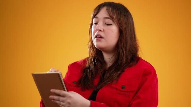Portrait of journalist woman making notes, writing down thoughts with pen in notepad notebook diary journal, to do list, good idea. Pretty brunette student girl isolated on yellow studio background