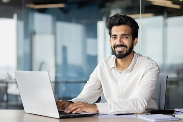 Fototapeta na wymiar Confident indian businessman with beard working on laptop at modern office desk