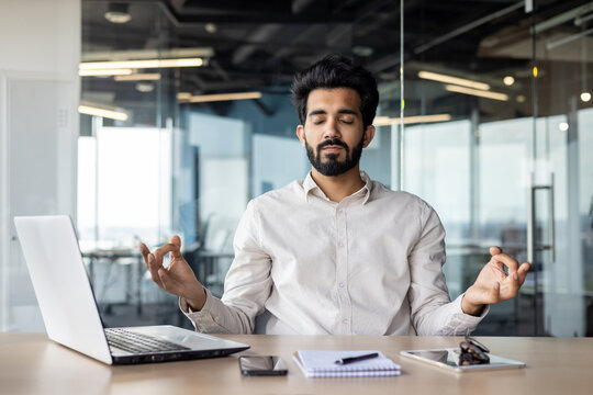 Indian businessman meditating at office desk with laptop for mindfulness