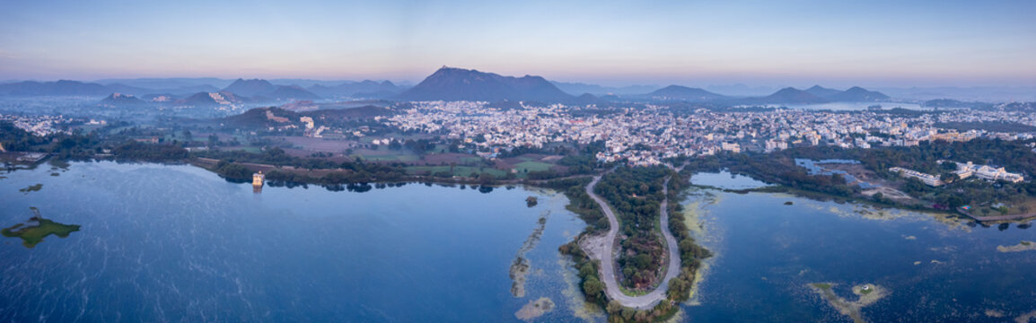 Panoramic aerial drone shot at dawn dusk with road loop extending into fateh sagar lake with aravalli hills in distance hidden in fog showing cityscape of Udaipur Rajasthan