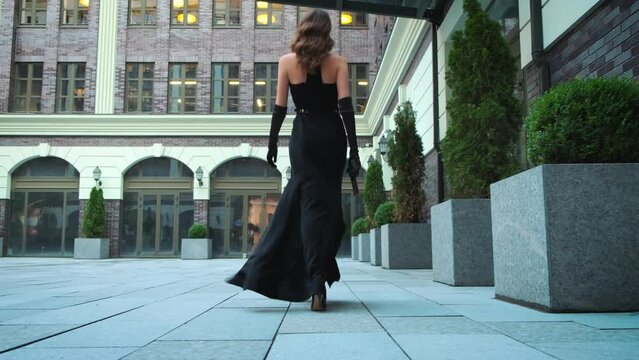 Happy Sexy fashion model woman posing walking down the street modern city back rear view. Elegant girl Long flowing loose light brown hair curl. Black evening gown prom dress. joy Beauty retro style