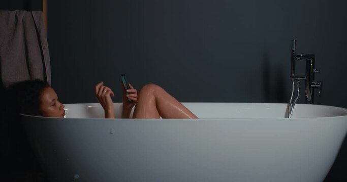 Side view beautiful young African American woman relaxing in modern glamour bath, using smart phone and taking video.