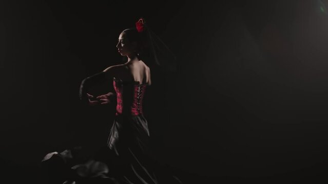 Woman dancing on black background under spotlights. Spanish dancer in red-black dress dancing elements of passionate flamenco.