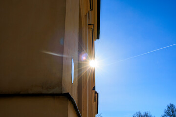 The sun shines over the edge of an old building under a blue sky with the vapour trail of an...