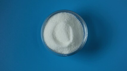 Glucono Delta-Lactone (GDL), also known as gluconolactone, organic white powder chemical formula (HOCH)3(HOCH2CH)CO2. Food additive E575 in glass dish on blue background. 4K video, rotating.