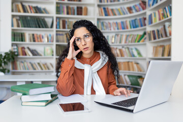 Irritated public library worker rolling eyes and leaning head on hand by desk with computer and...