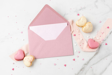Blank paper card and open retro pink envelope with macarons macaroons in heart shape and sprinkles on white marble background. Mockup birthday or valentines day greeting card