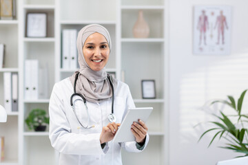 Beautiful arabic medical woman standing in workspace with digital tablet in hands. Confident female...